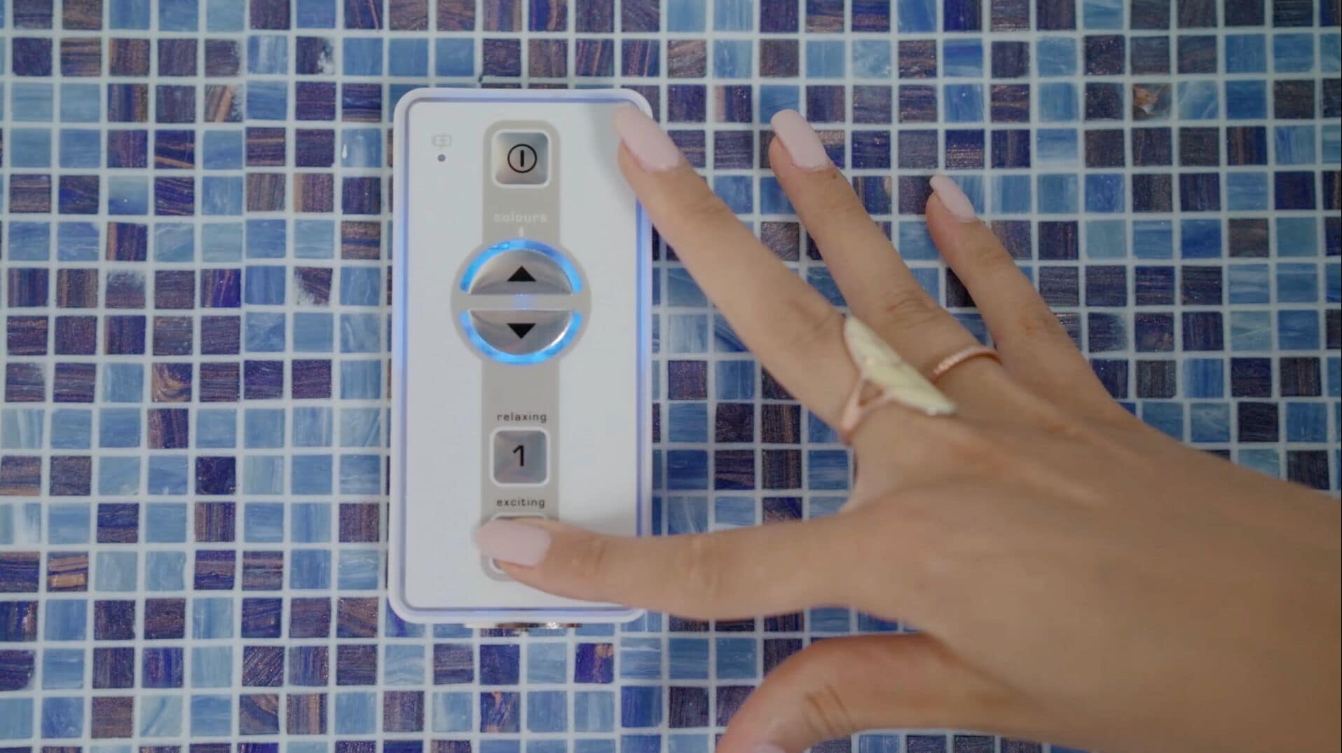 A hand holding a white device with a circular dial and a button labeled "1" on a tiled wall.