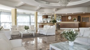 A spacious lobby with white sofas, a glass coffee table, and a reception area with wooden paneling and a chandelier. - Hotel Meeting (Riccione)