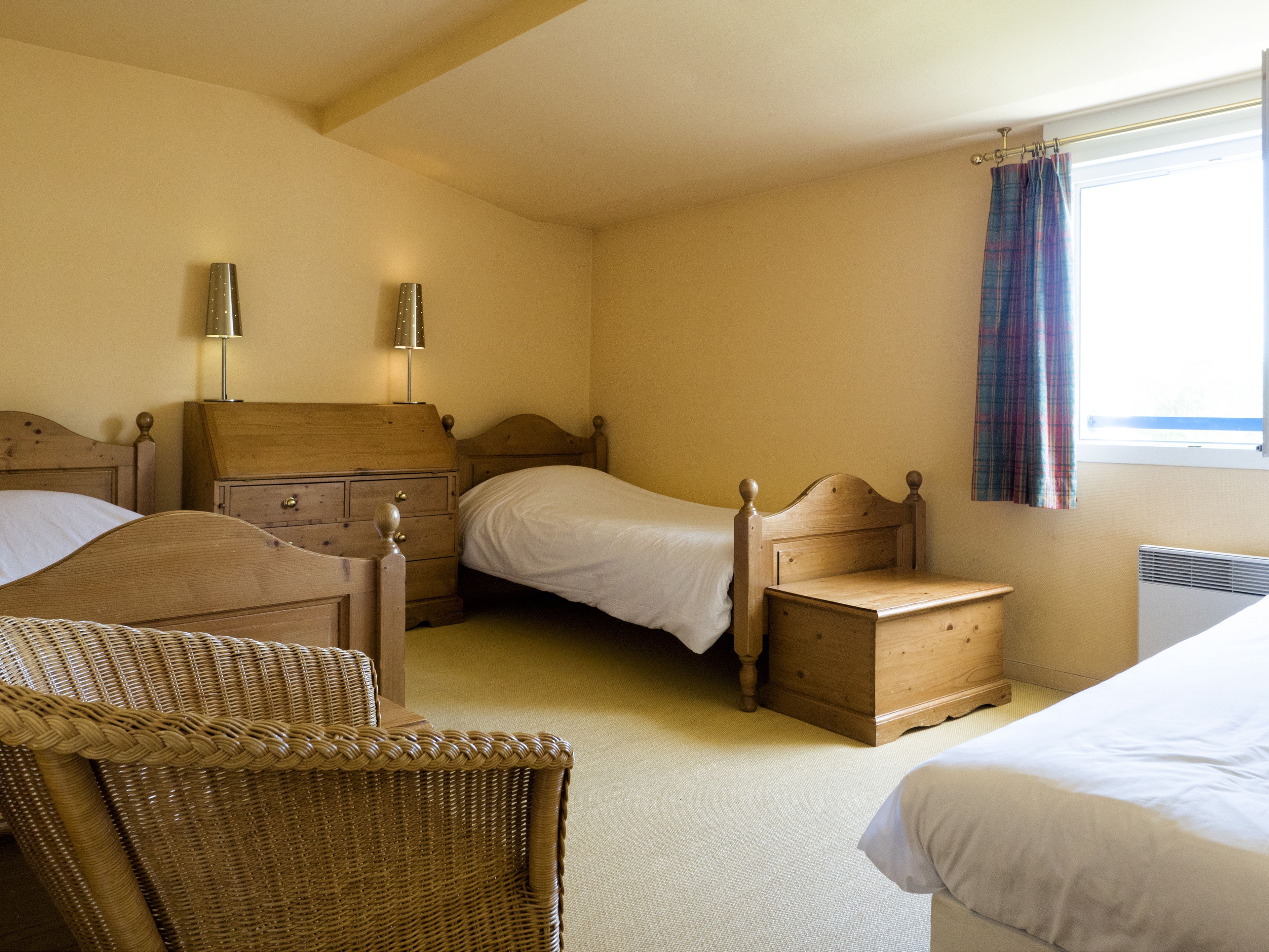 A bedroom with two wooden beds, a wicker chair, a small wooden chest, and a nightstand with a lamp.