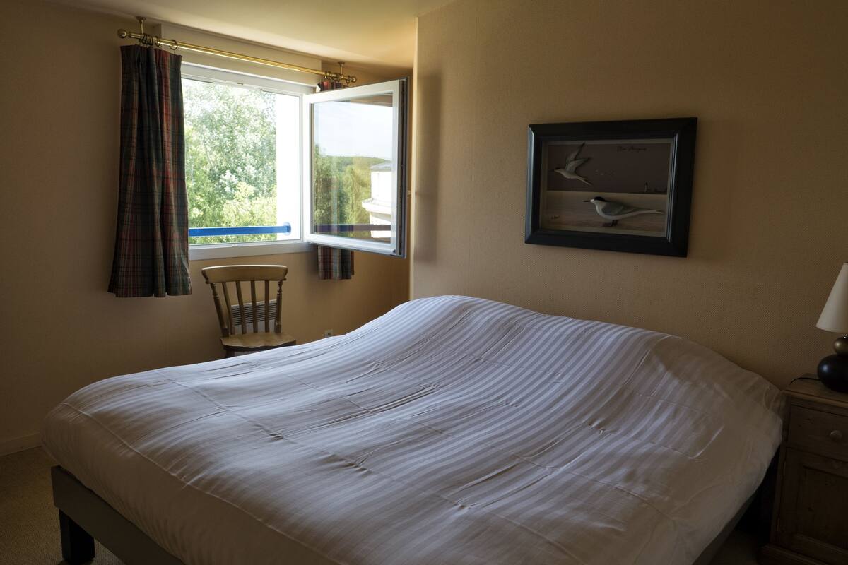 A neatly made bed in a room with a window, a chair, and a framed picture on the wall.