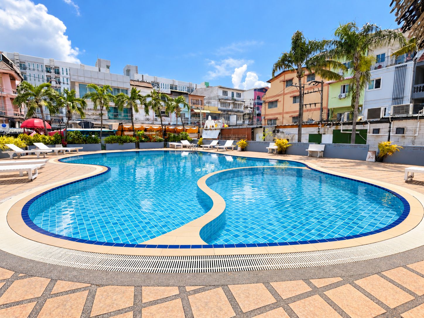 A swimming pool with tiled edges, surrounded by lounge chairs and palm trees, with buildings in the background.