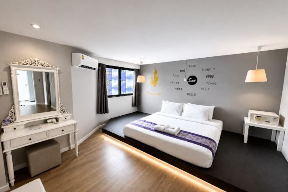 A modern hotel room with a large bed, a vanity with a mirror, a desk, and a wall-mounted air conditioner.