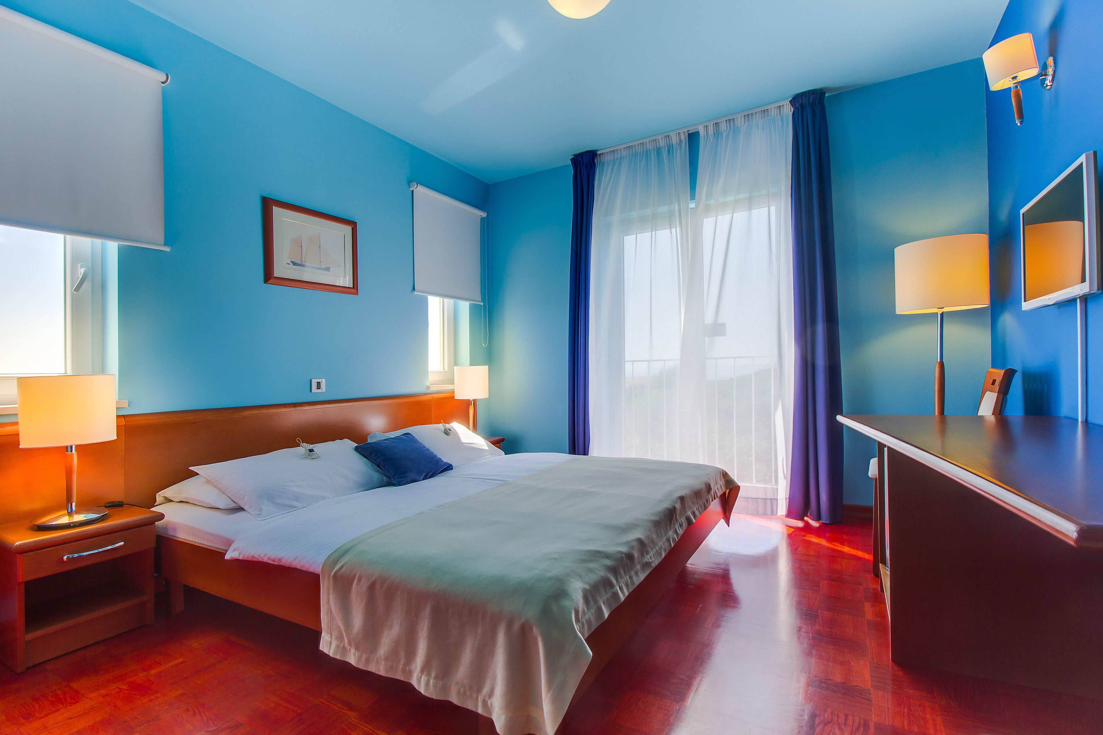 A hotel room with a large bed, a desk, and a chair. There is a window with curtains, a picture on the wall, and a lamp on the desk.