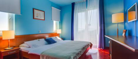 A hotel room with a large bed, a desk, and a chair. There is a window with curtains, a picture on the wall, and a lamp on the desk.