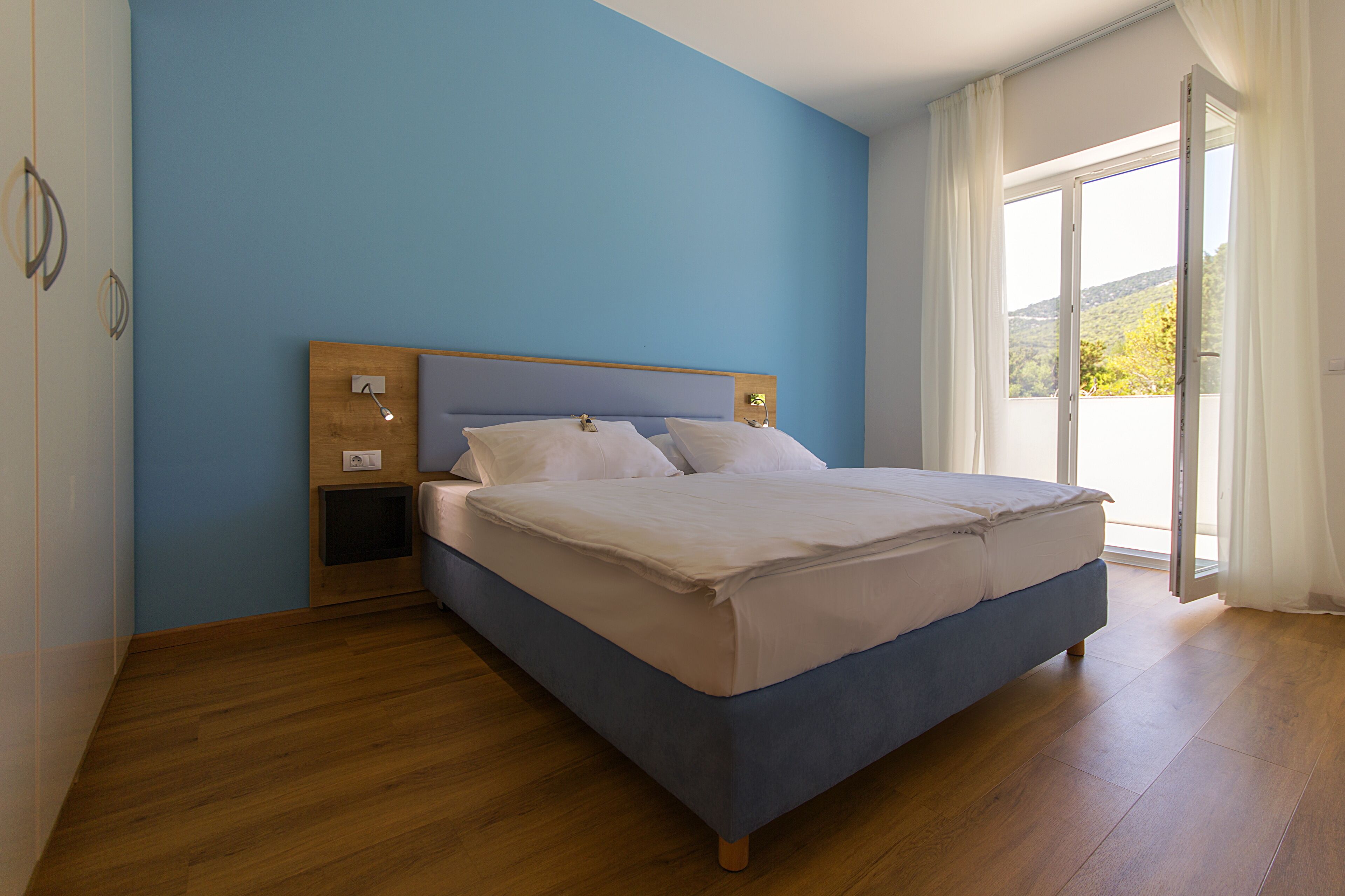 Quarto com cama, cabeceira de madeira, mesas de cabeceira e uma janela grande com cortinas.