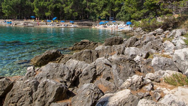 Una costa rocosa con agua turquesa cristalina, una playa con sombrillas y un fondo boscoso.