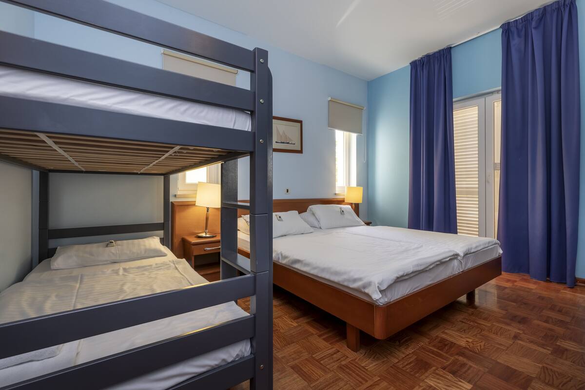 A bunk bed room with a wooden floor, a single bed, a nightstand, a lamp, and a window with curtains.