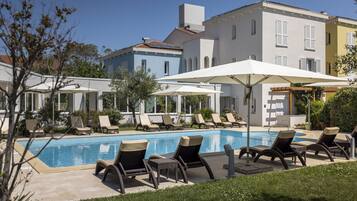 Ein Poolbereich mit Liegestühlen und Sonnenschirmen, ein klares, blaues Schwimmbecken und ein mehrstöckiges Gebäude mit Balkonen.