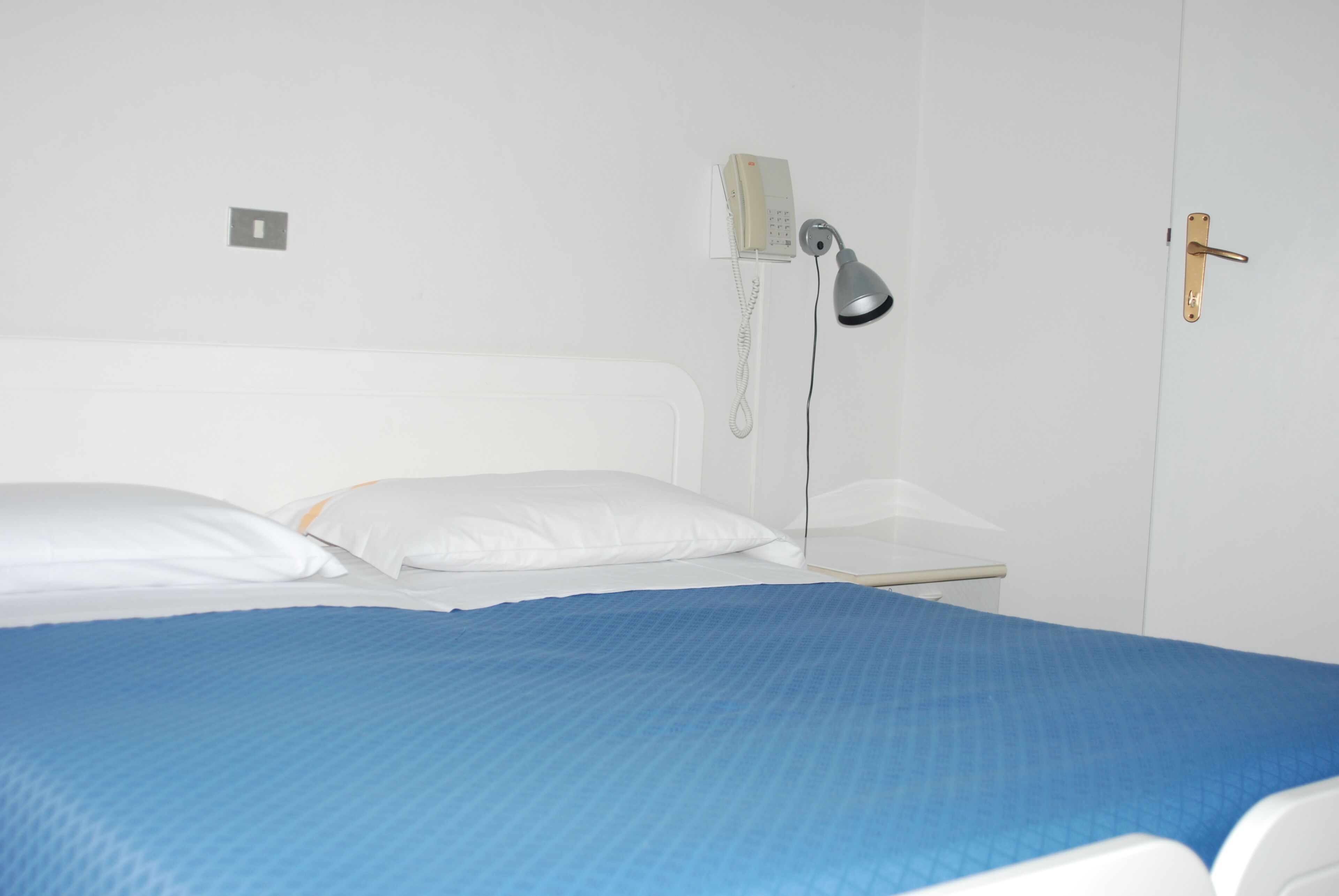 Un letto con una coperta blu, un telefono sul comodino e una porta con maniglia.