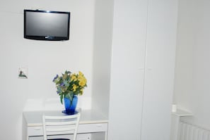 A white desk with a blue vase of flowers, a white chair, and a mounted television above. - Hotel Tuscolano (Bologna)