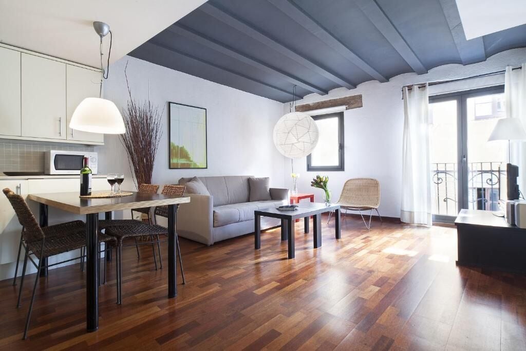Een moderne woonkamer met een eethoek, een bank, een salontafel en een stoel. Er is een raam met uitzicht, een lamp en een schilderij aan de muur.