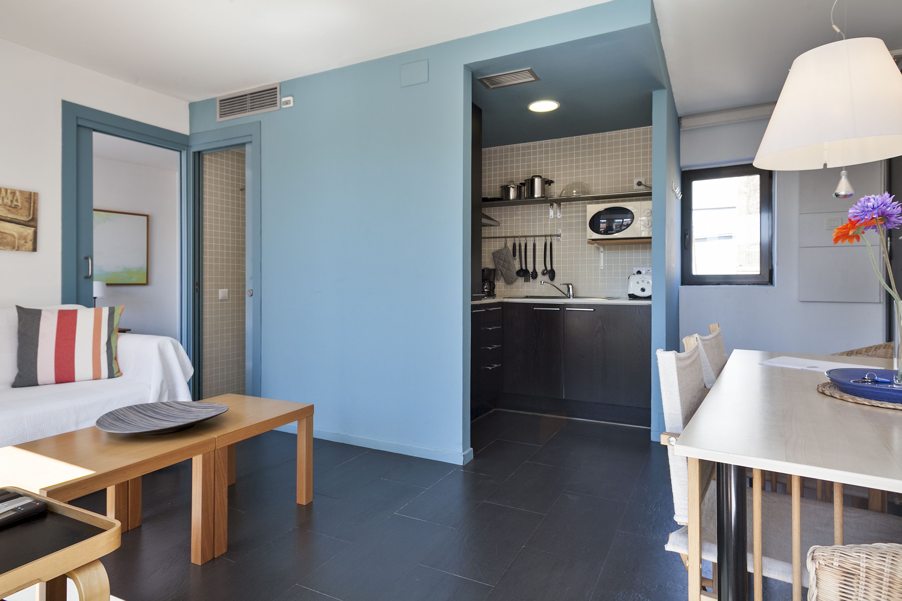A modern kitchen with dark tiles, a dining area with a wooden table, and a living space with a sofa and a painting.
