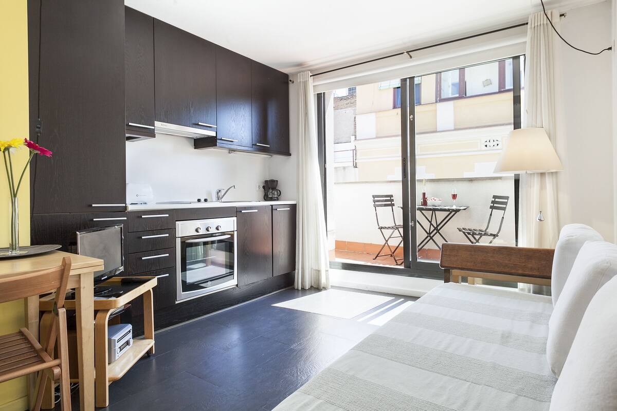 Una cocina compacta con armarios oscuros, un rincón comedor con mesa y sillas, y un sofá.