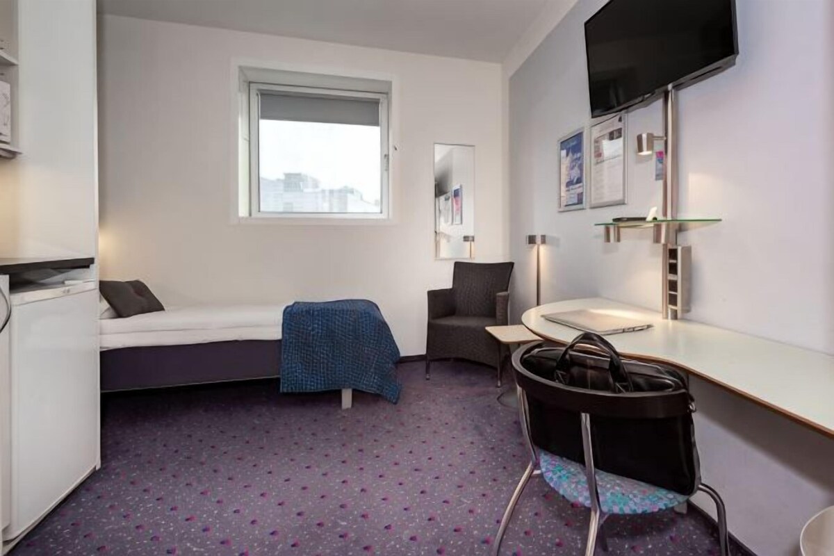 A compact hotel room with a bed, a desk, a chair, a small table, a TV, and a window.