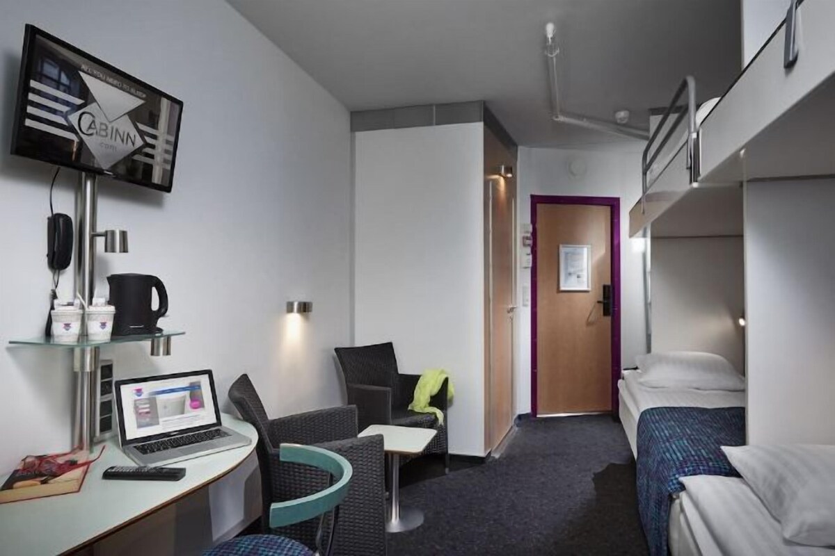 A hotel room with a bed, a desk with a laptop, a chair, a television, and a door leading to another room.