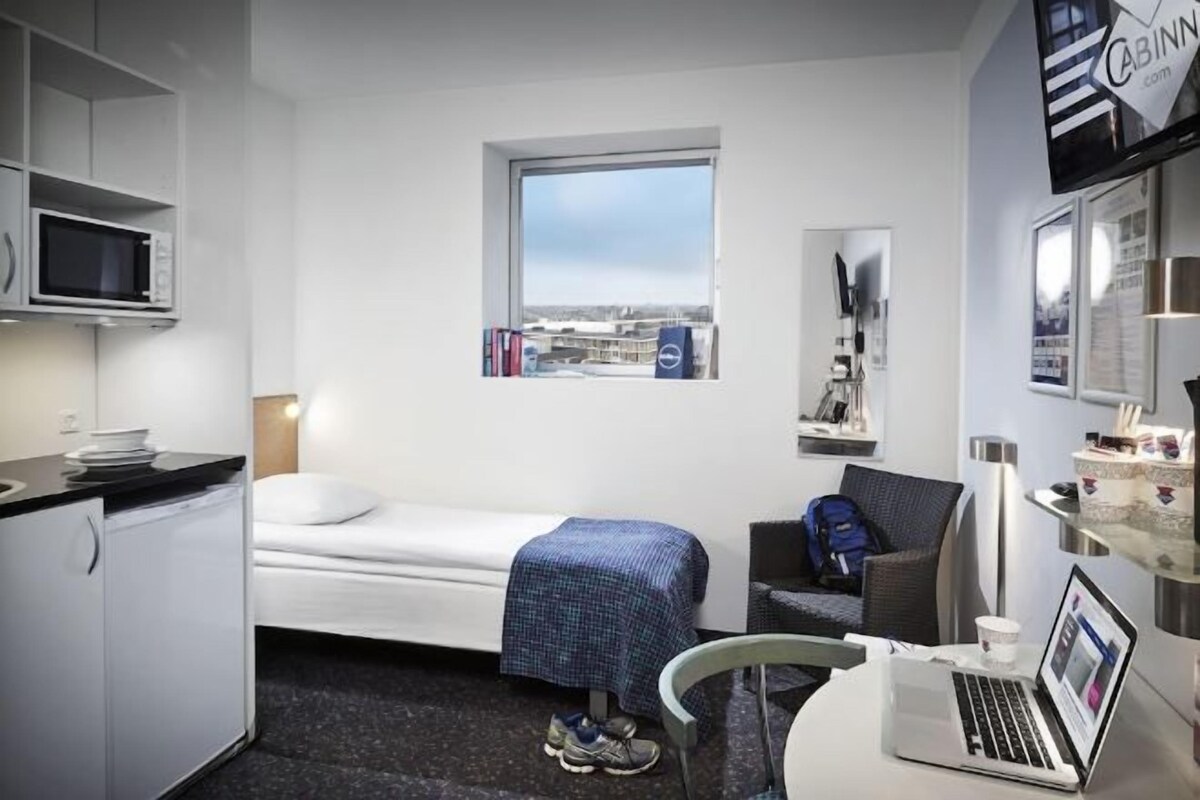 A compact hotel room with a bed, a desk with a laptop, a chair, a microwave, and a window with a view.