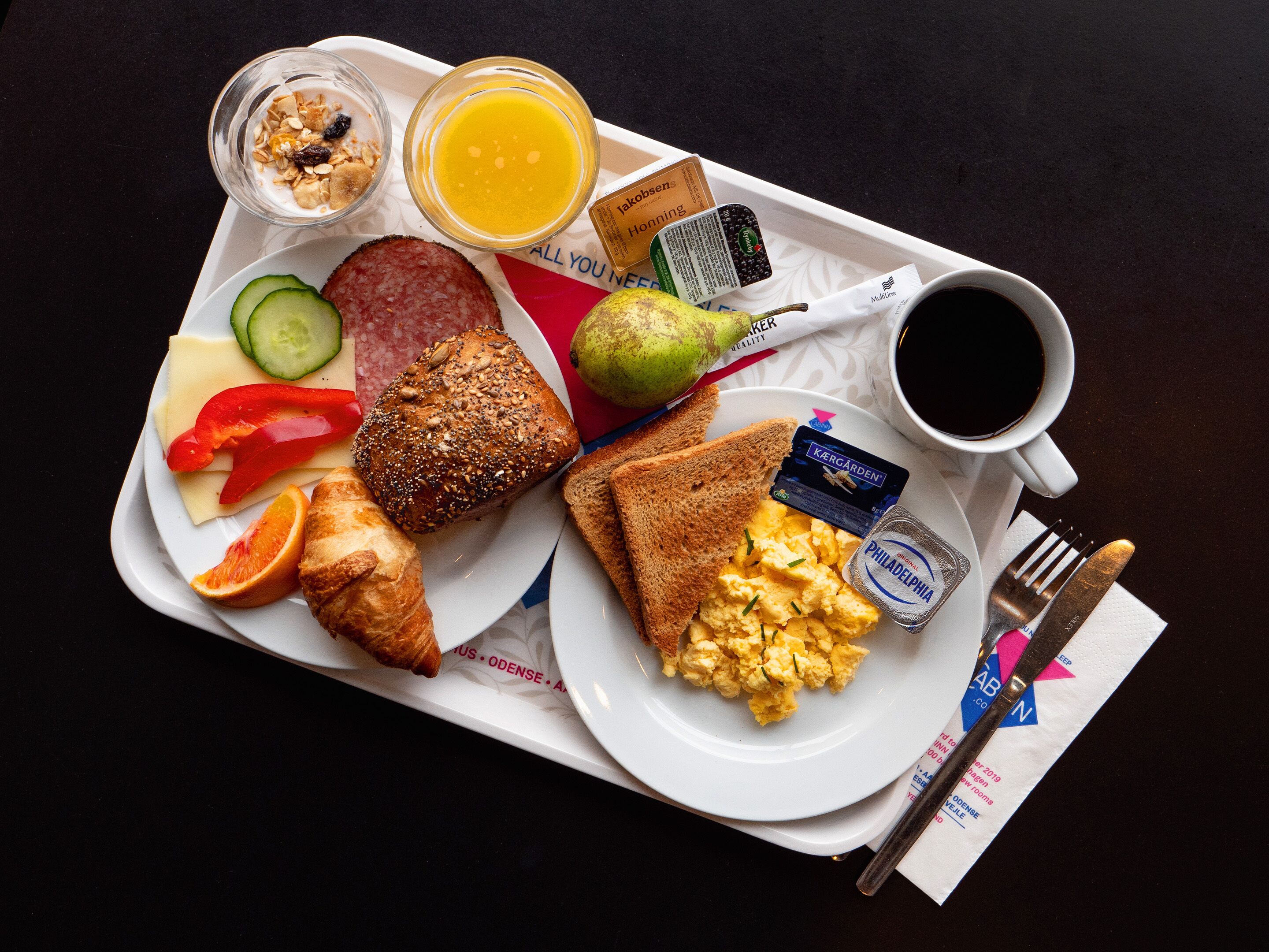 A breakfast tray with toast, scrambled eggs, a croissant, a slice of cheese, a red bell pepper, a cucumber, a sausage, a pear, a glass of orange juice, a cup of coffee, and a small container of jam.