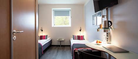 Quarto de hotel compacto com cama, mesa pequena, escrivaninha com cadeira, televisão e janela.