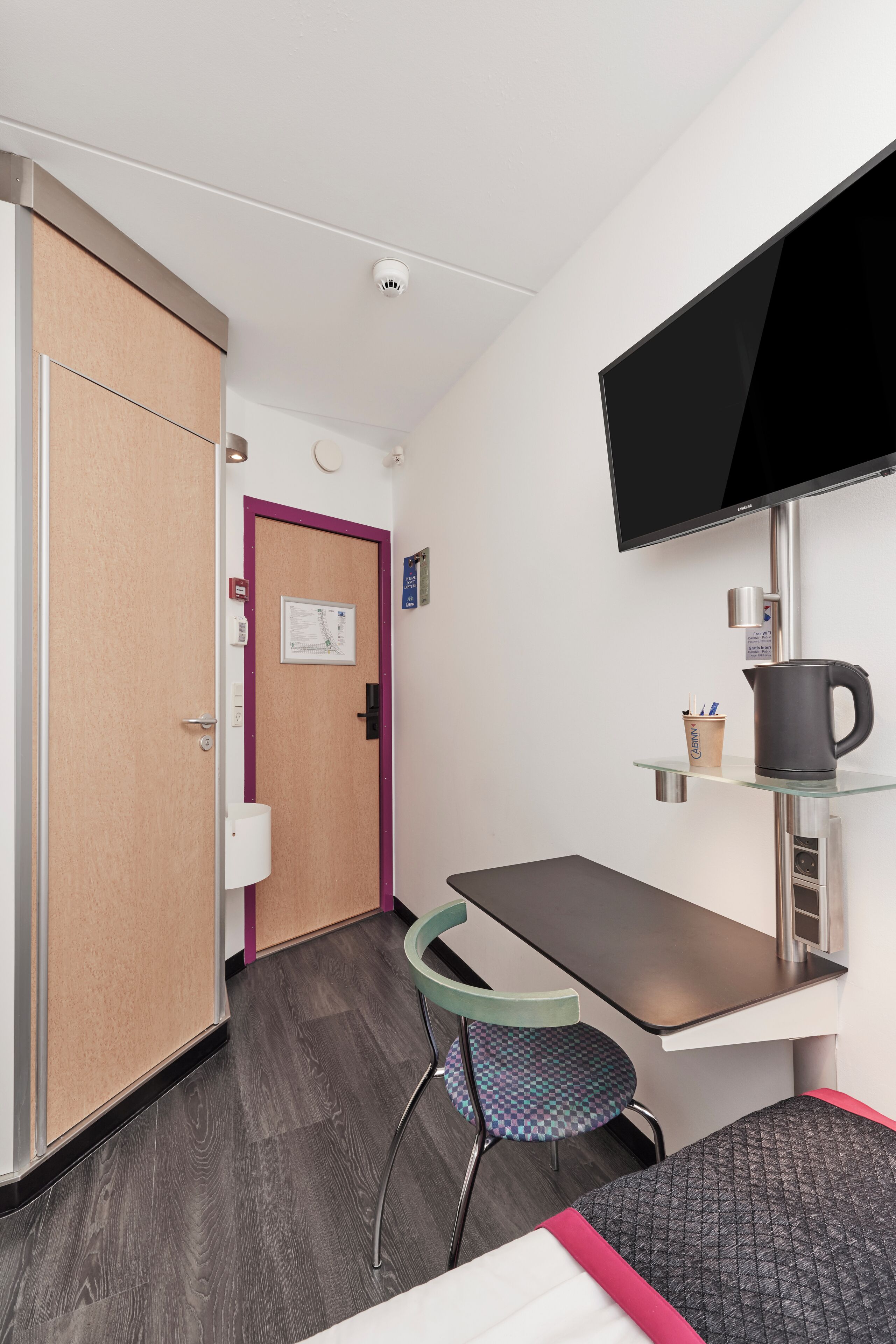 A compact hotel room with a single bed, a small desk, a chair, a television mounted on the wall, and a door leading to another room.