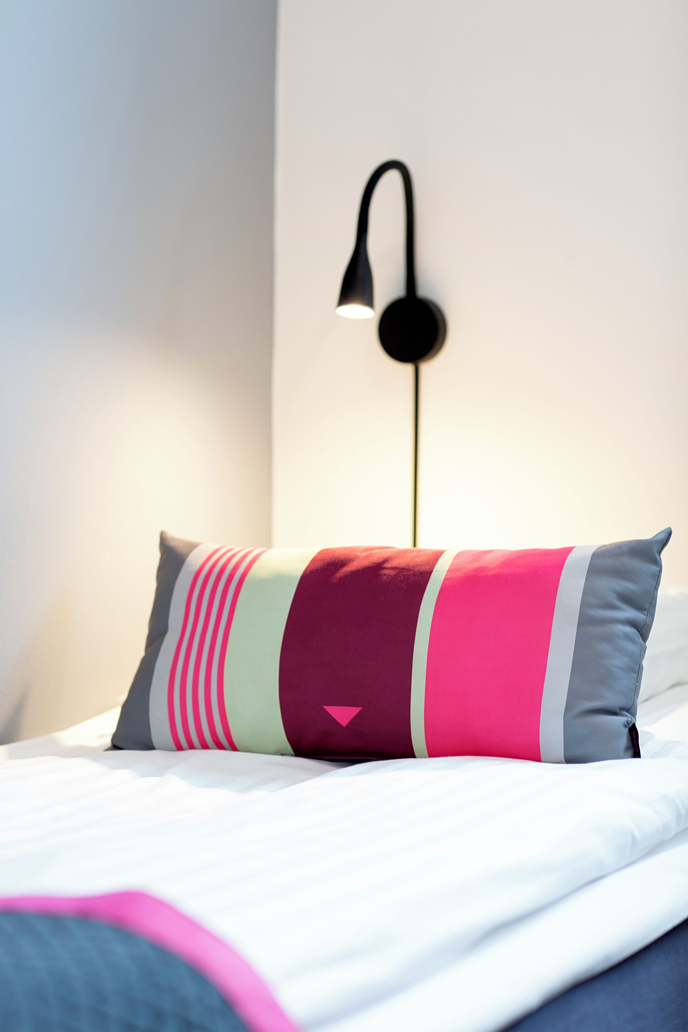Una cama con un cojín de colores y una lámpara negra fijada a la pared.
