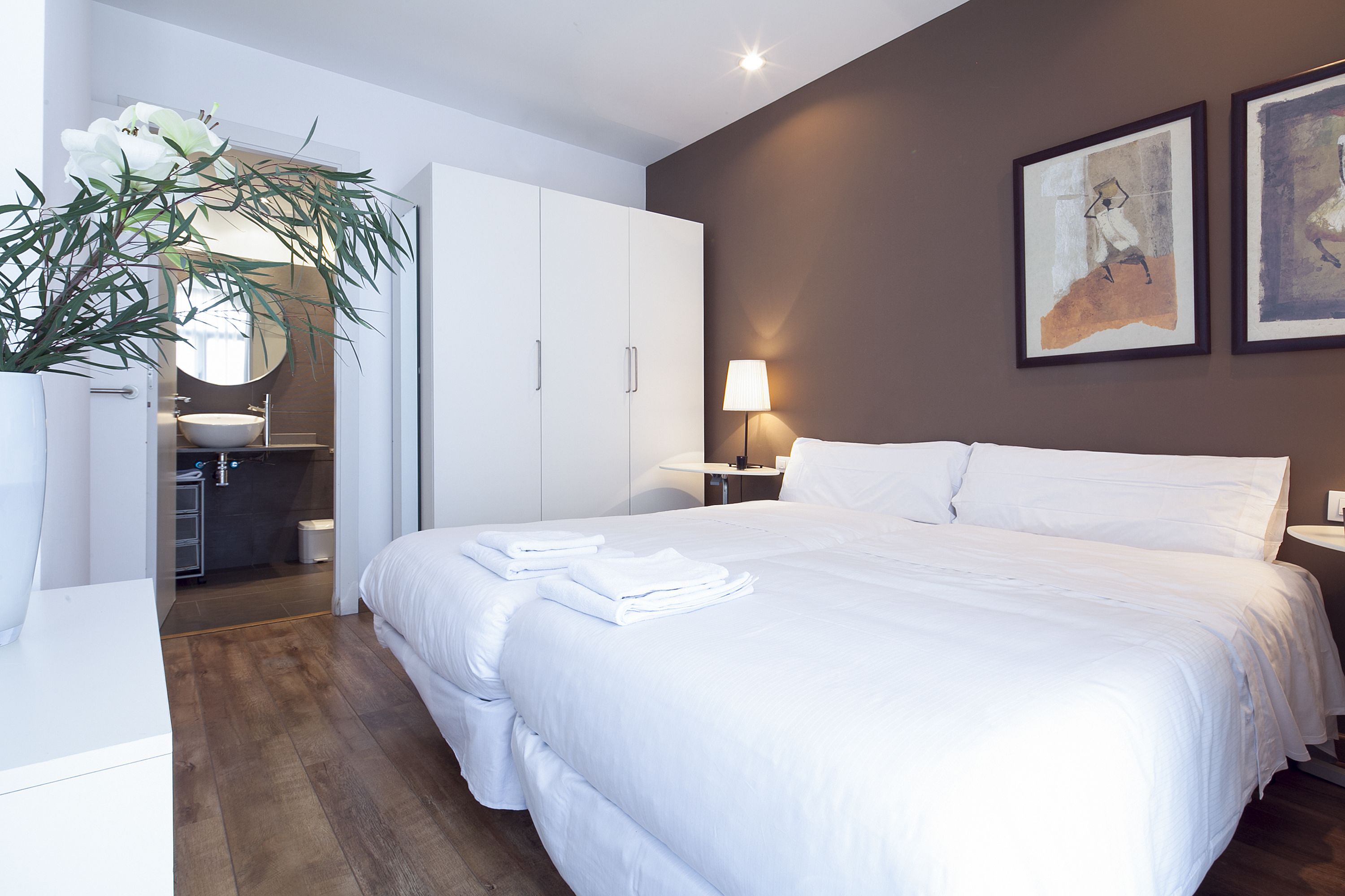 Quarto de hotel com uma cama grande, um armário branco, banheiro com pia e espelho, e duas obras de arte emolduradas na parede.