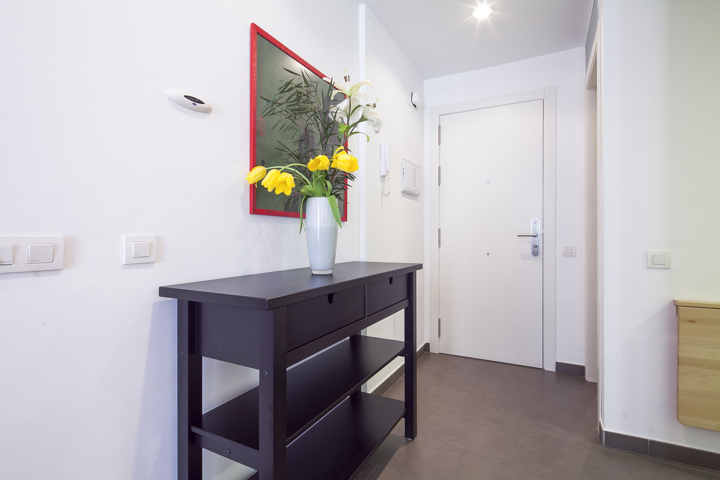 Un couloir avec une console noire, un vase contenant des fleurs jaunes, un miroir à cadre rouge et une porte blanche.