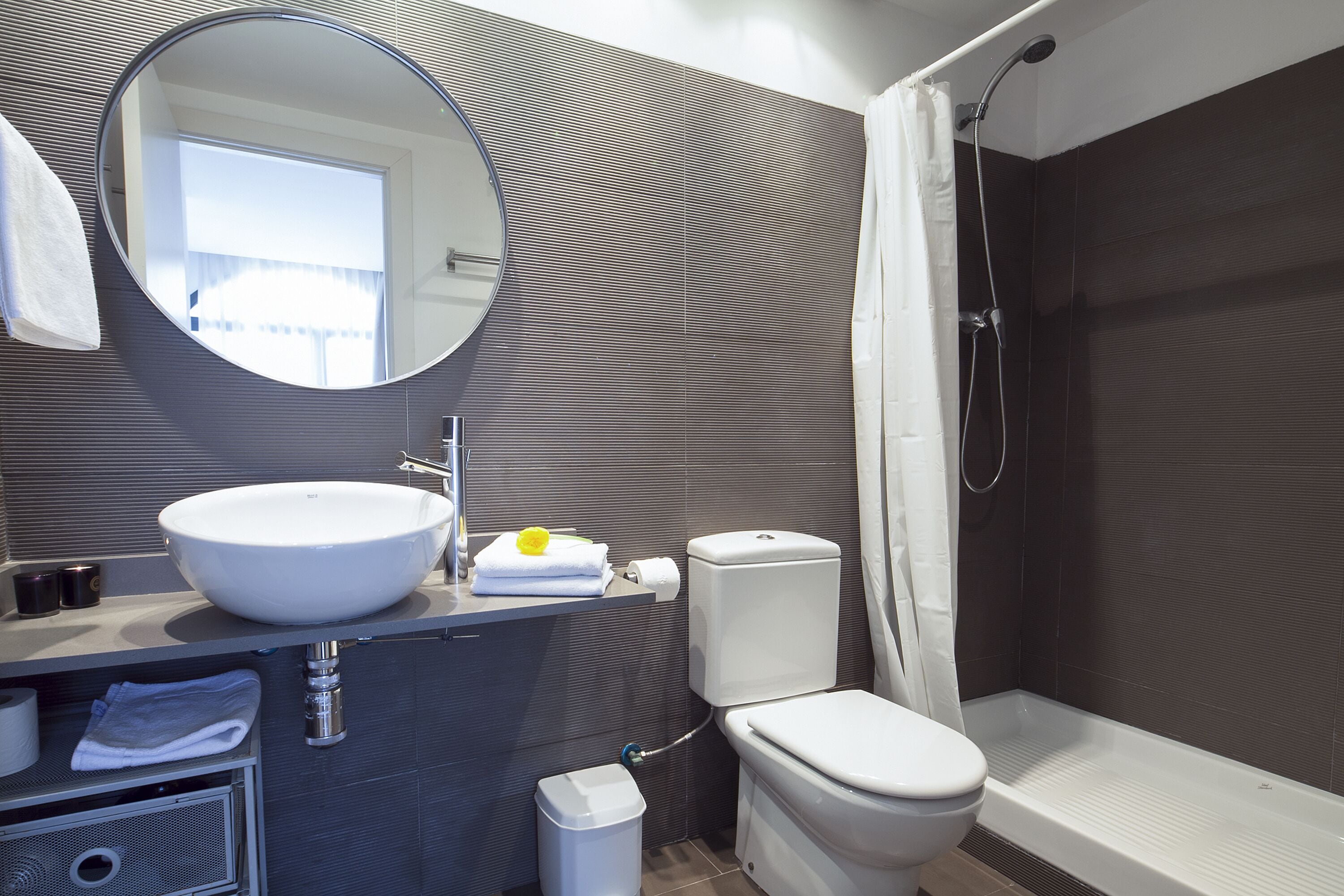 A modern bathroom with a round mirror, white sink, toilet, and shower.
