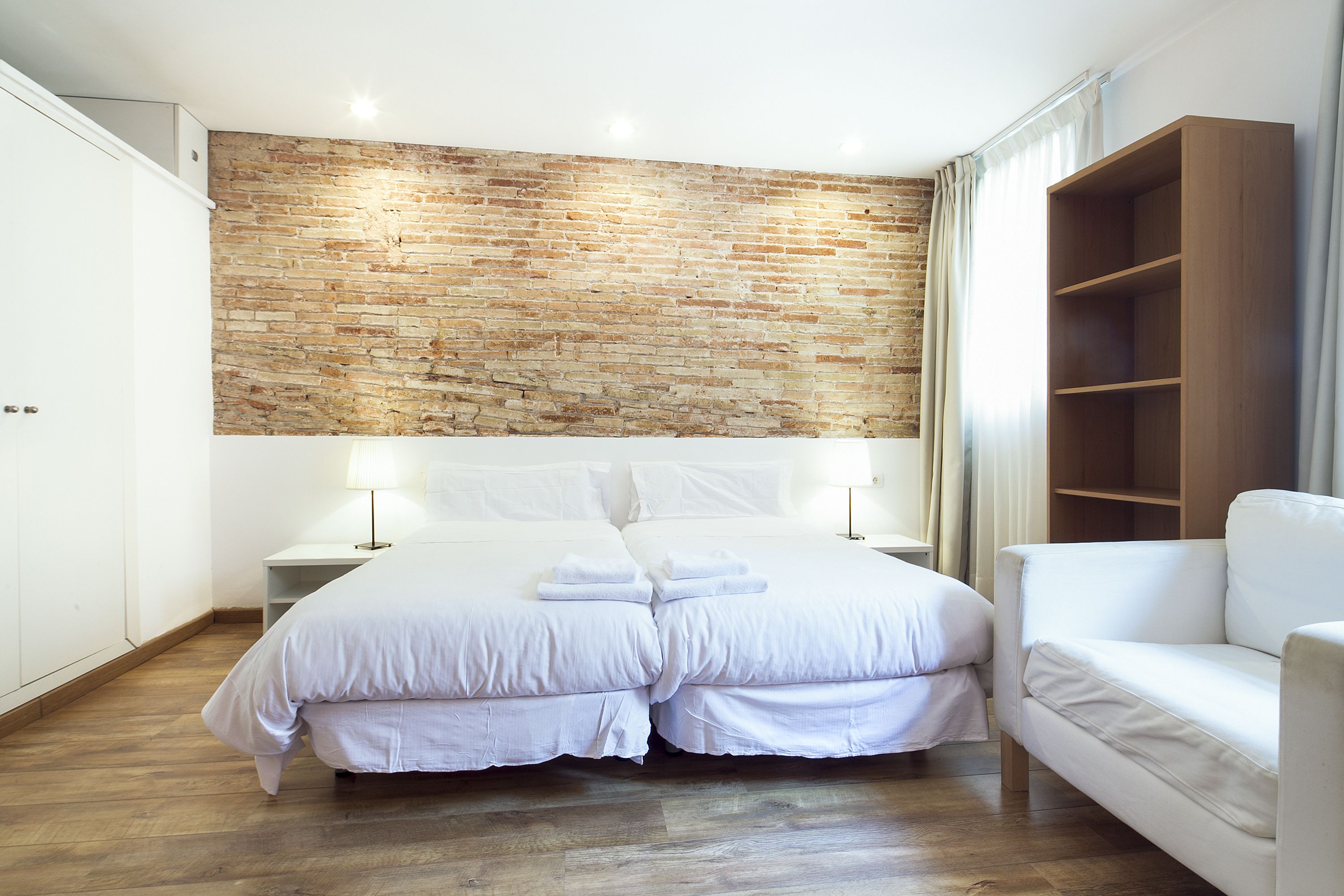 Une chambre avec un mur en briques, un lit avec une literie blanche, une table de chevet, une chaise blanche et une étagère en bois.