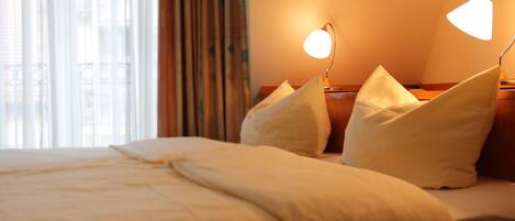 A hotel room with a neatly made bed, two bedside lamps, and a view of the outdoors through large windows.