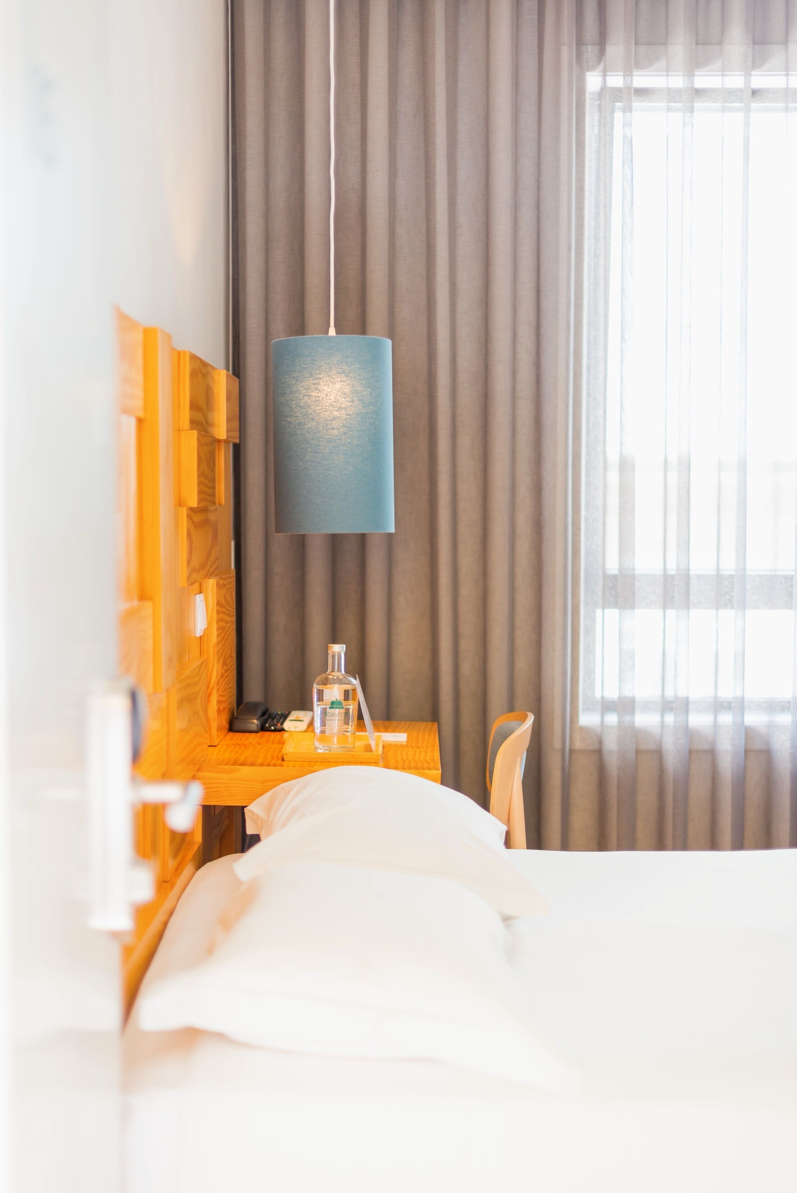 Habitación de hotel con una cama, una mesita de noche de madera, una lámpara, una botella y una silla.