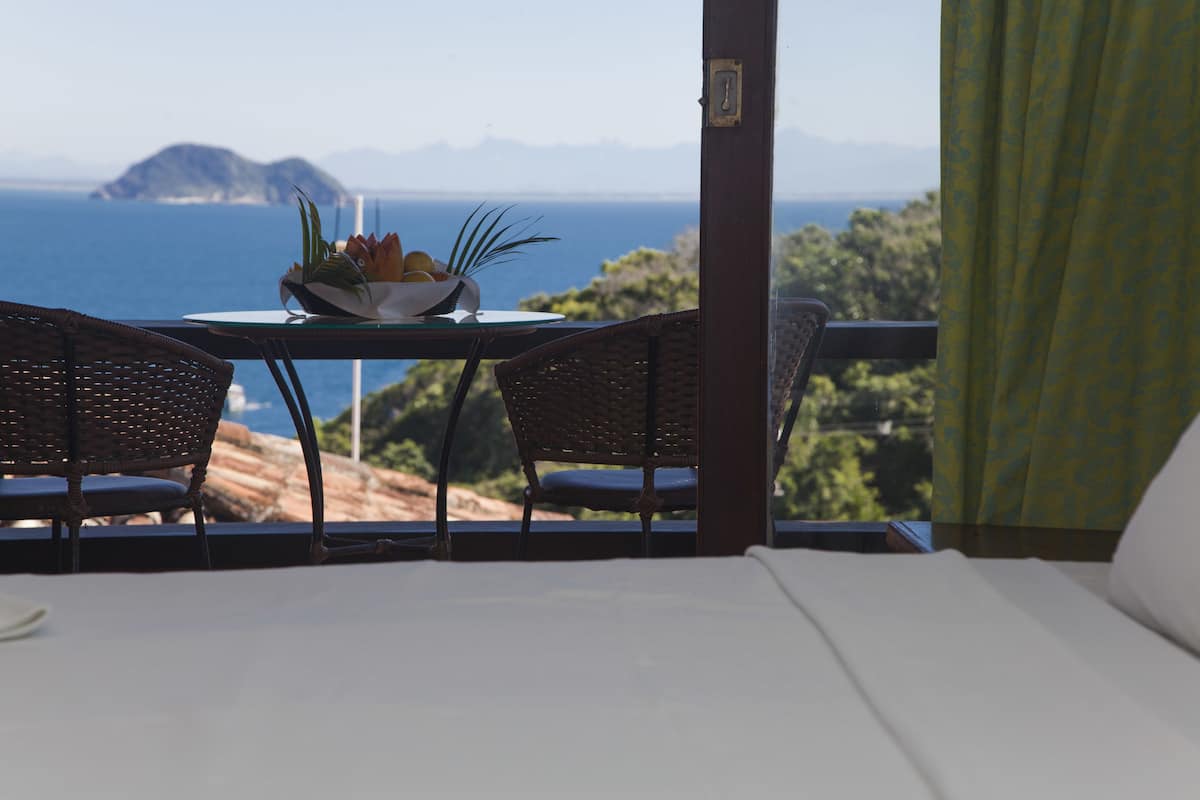 A balcony with wicker chairs, a table with a fruit bowl, and a view of the sea and mountains.
