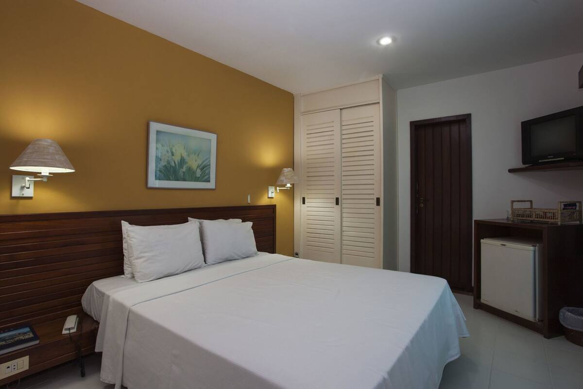 A hotel room with a bed, bedside lamps, a painting, a wardrobe, a television, and a small refrigerator.