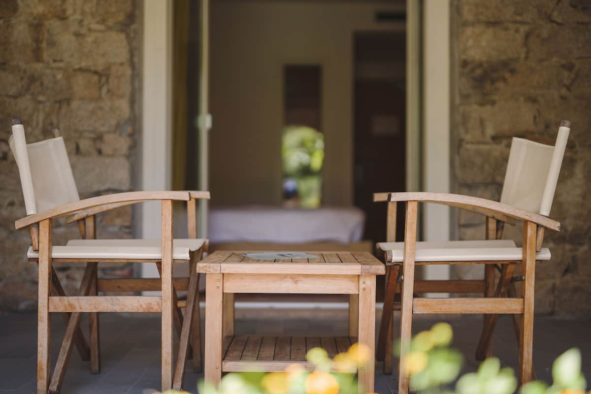 Two wooden chairs and a small table in a stone-walled room with a bed in the background.