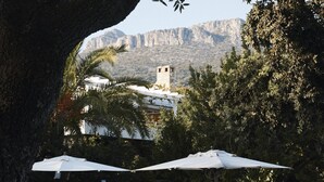 Zona de piscina con sillones de descanso y sombrillas, un muro de piedra y una vista de una montaña y un edificio al fondo.