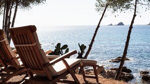 Una terraza de madera con dos sillones de descanso frente a un mar tranquilo, rodeada de cactus y rocas.
