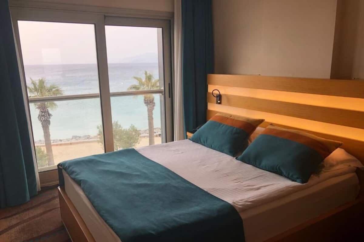 Ein Hotelzimmer mit Bett, Nachttisch, Fenster mit Meerblick und Vorhang.