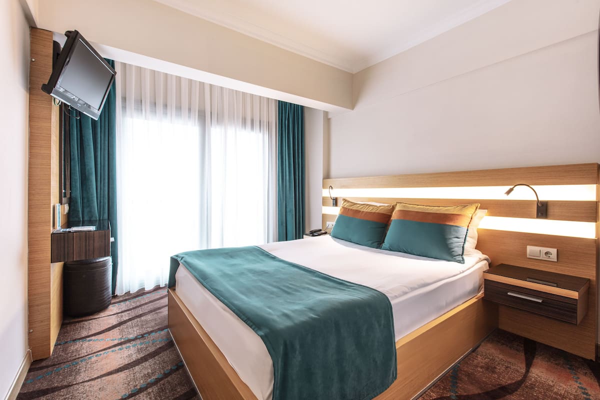 Ein Hotelzimmer mit Bett, Nachttisch, Lampe, Fernseher und einem Fenster mit Vorhängen.