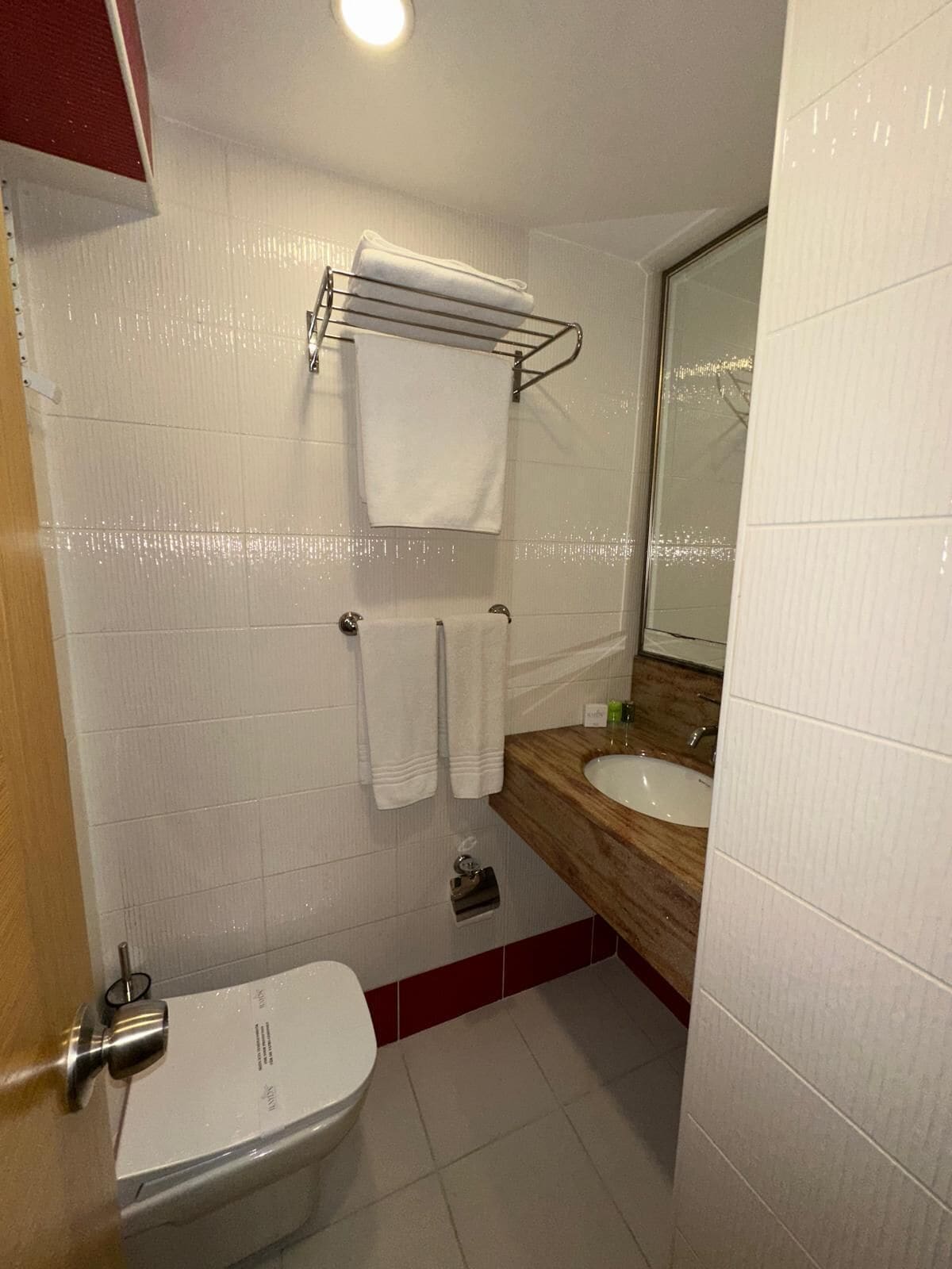 Un baño con inodoro blanco, tocador de madera con lavamanos y espejo. Hay dos toallas colgadas en un perchero.