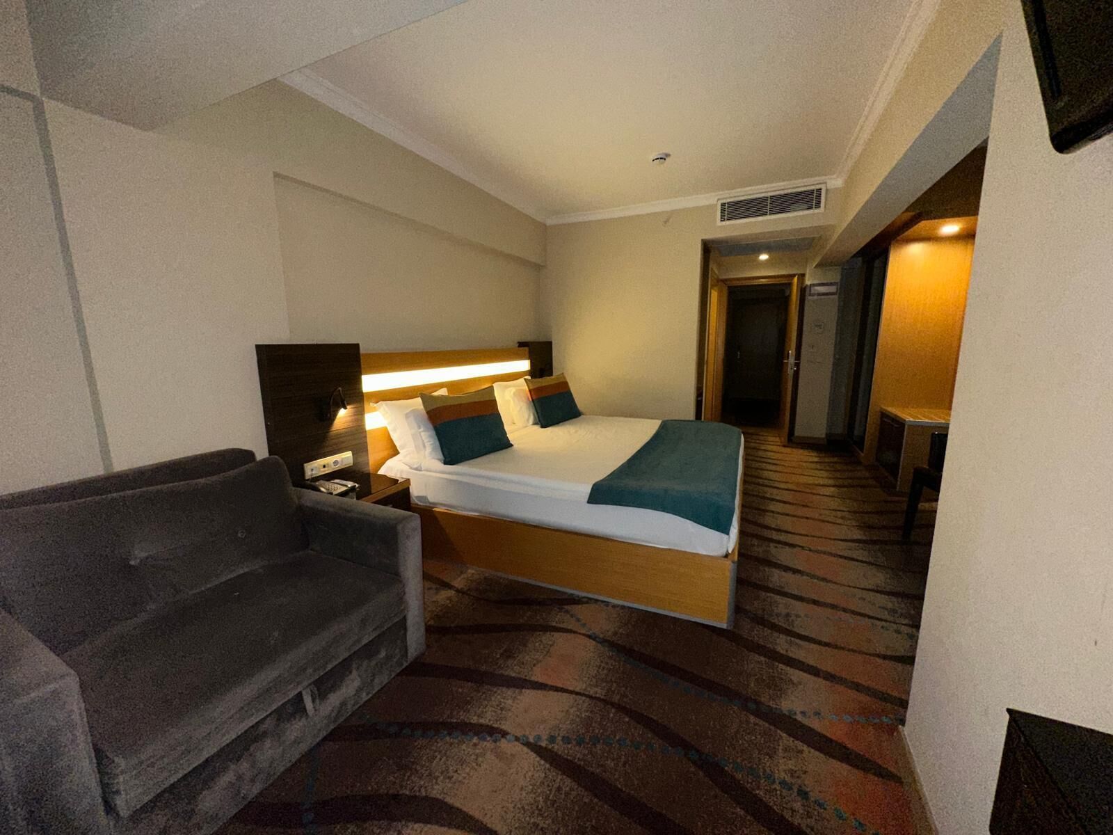 A hotel room with a bed, a bedside table, a lamp, a sofa, and a door leading to another room.