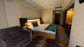 A hotel room with a bed, a bedside table, a lamp, a sofa, and a door leading to another room.