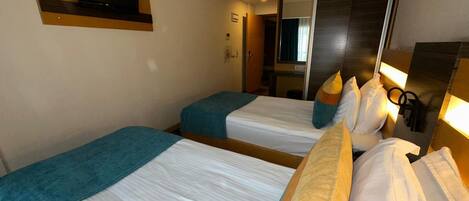 Habitación de hotel con dos camas, cada una con una manta azul y ropa de cama blanca. Hay un espejo grande en la pared, un televisor montado sobre las camas y una puerta que da a un baño.
