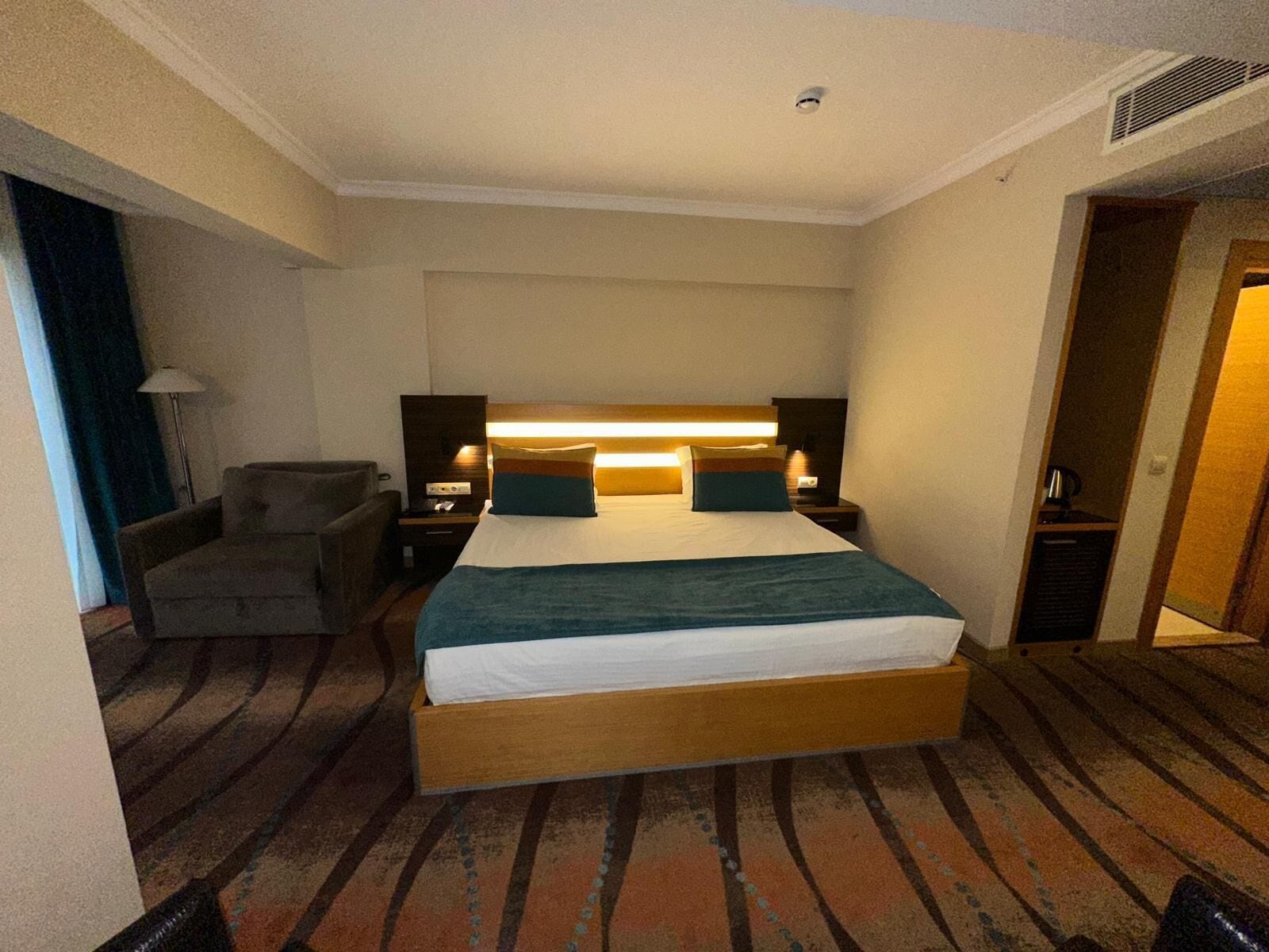 A hotel room with a large bed, a chair, a desk, and a nightstand with a lamp.