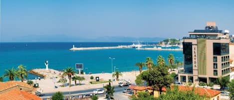 Un paisaje urbano costero con un hotel destacado llamado "HOTEL ILAYDA", de diseño moderno, ubicado cerca de un puerto deportivo con barcos y vistas despejadas al mar.