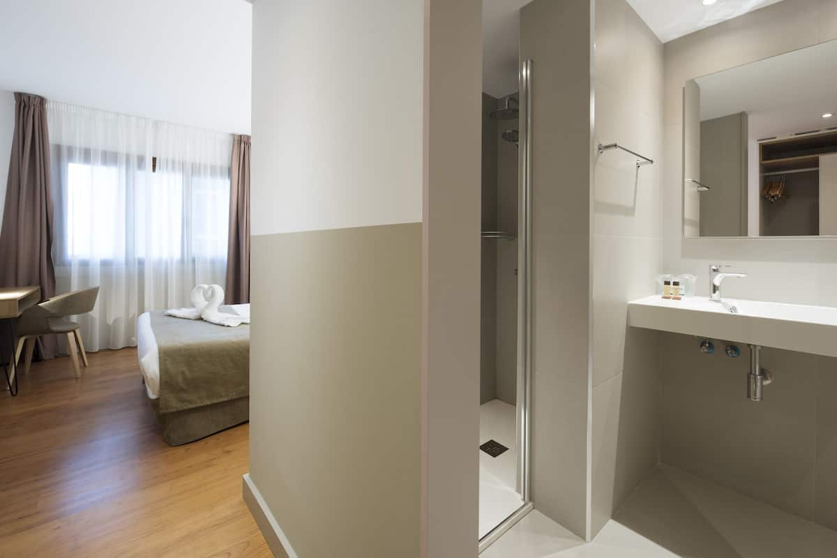 Ein modernes Hotelzimmer mit Bett, Schreibtisch, Dusche und Bad mit Waschbecken und Spiegel.