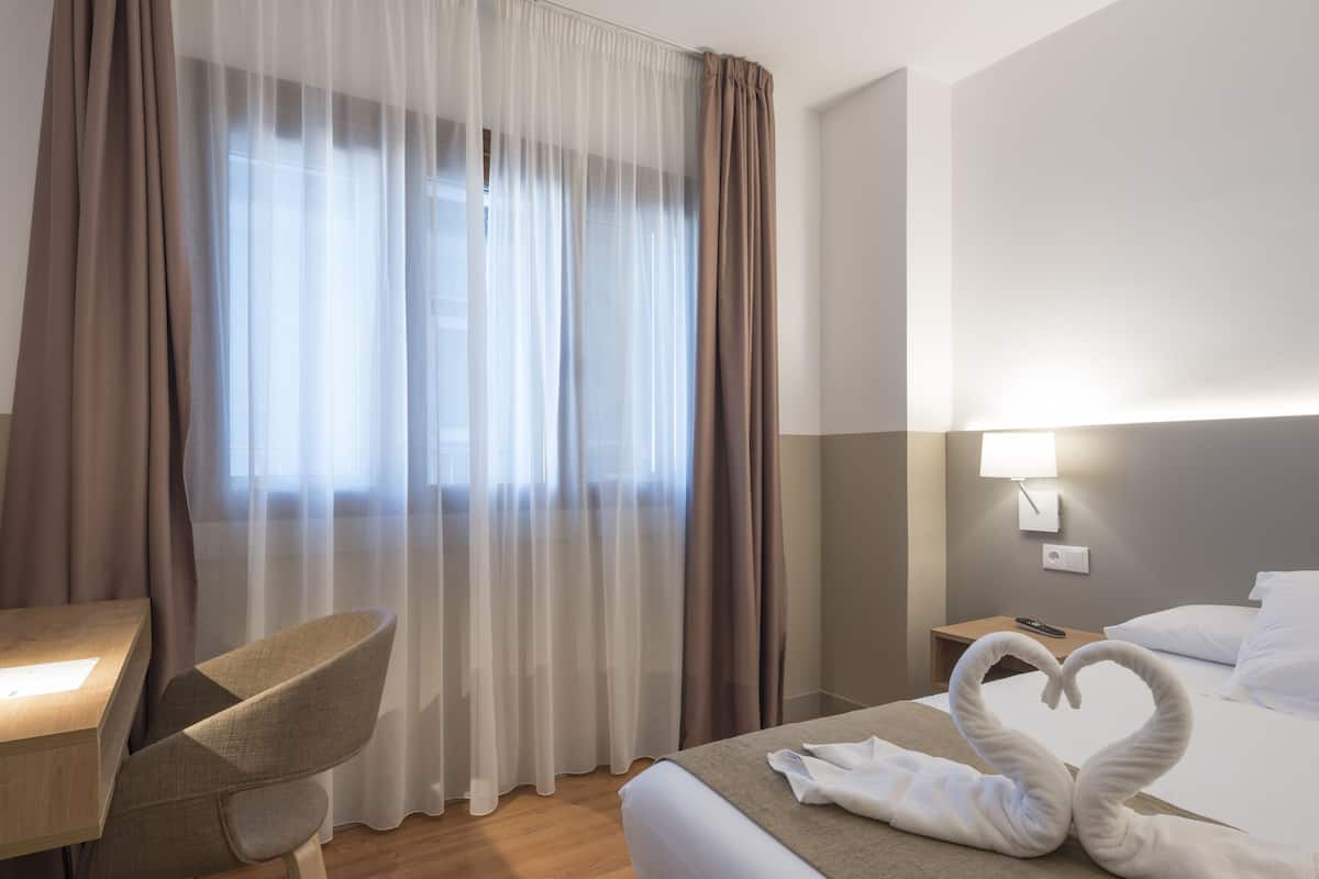 Ein Hotelzimmer mit Bett, Schreibtisch, Stuhl und einem Fenster mit Vorhängen.