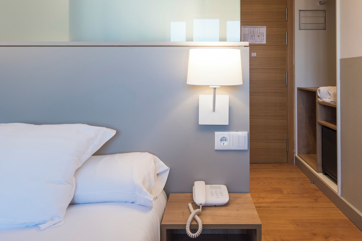 A hotel room with a bed, a bedside table with a phone, a wall-mounted lamp, and a wooden wardrobe.