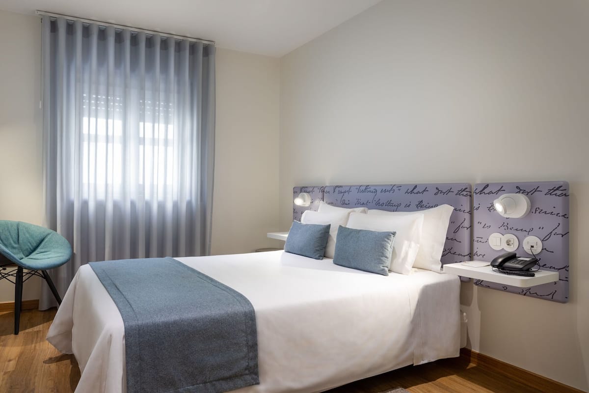 A neatly made bed with a white and grey bedspread, two pillows, and a bedside table with a lamp and phone.