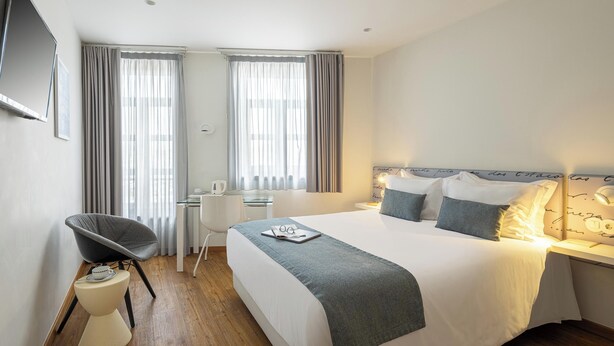 Una habitación de hotel moderna con cama, mesita de noche, silla, escritorio y cortinas.