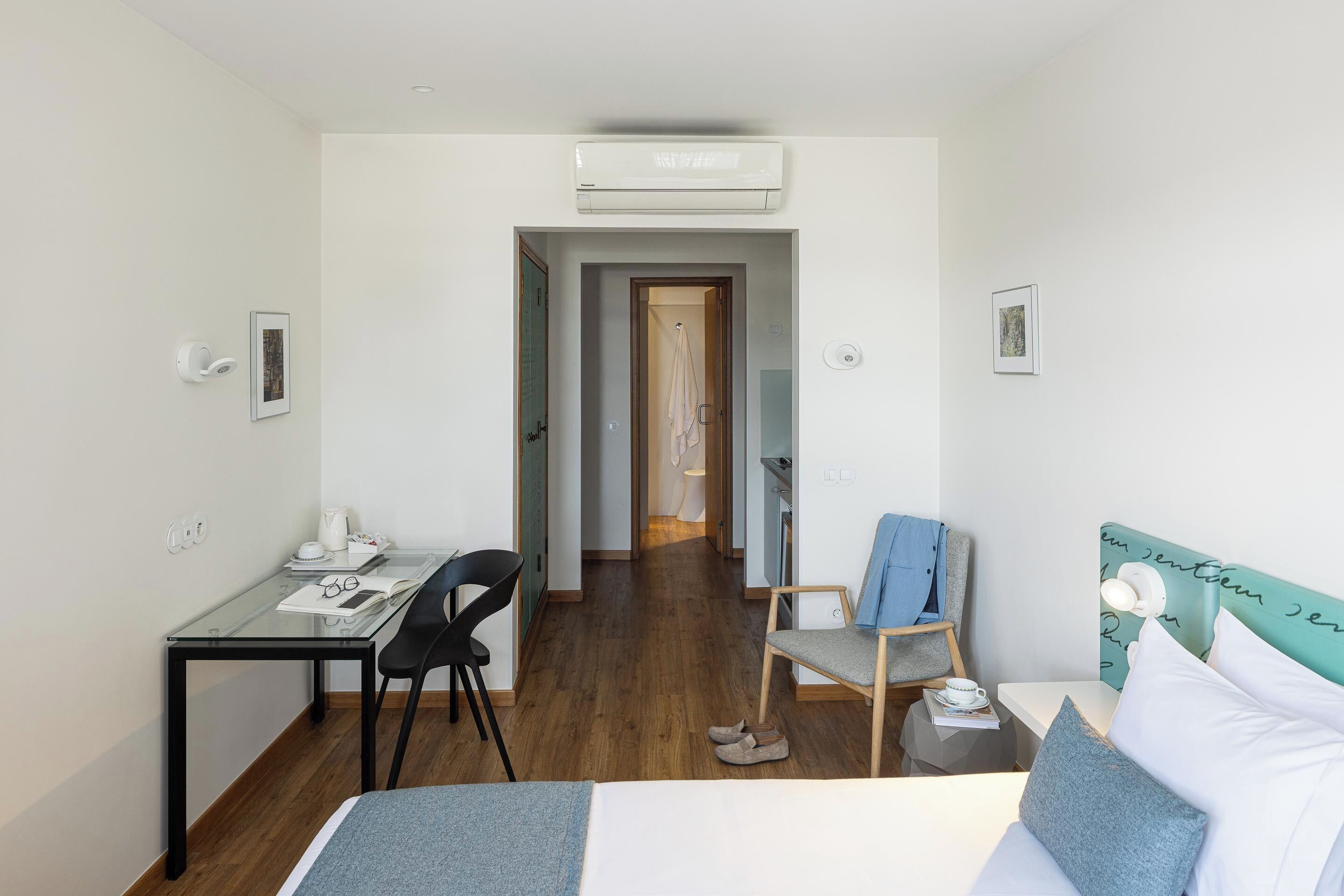 Una habitación de hotel moderna con un escritorio de vidrio, una cama con cabecera color turquesa, suelo de madera y un pasillo que conduce a un baño.