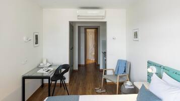 Una habitación de hotel moderna con un escritorio de vidrio, una cama con cabecera color turquesa, suelo de madera y un pasillo que conduce a un baño.